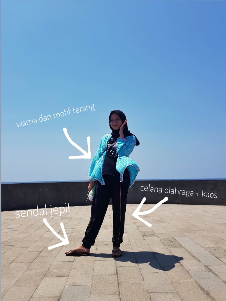 Woman wearing light blue cardigan, black outfit, and sandals in front of sea and sky